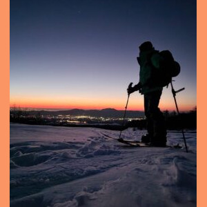 Imagem de Daniel Tamaki com roupas e acessórios de práticas de ski, ele está parado sobre a neve e ao fundo um lindo pôr do sol Daniel Tamaki, guia no Japão, com equipamento de esqui sobre a neve em um pôr do sol, representando a experiência de esqui e snowboard no Japão. Ideal para quem busca um guia para esportes de inverno em montanhas japonesas.