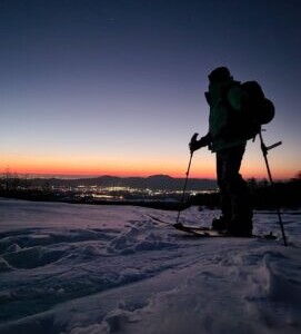 Imagem de Daniel Tamaki com roupas e acessórios de práticas de ski, ele está parado sobre a neve e ao fundo um lindo pôr do sol Daniel Tamaki, guia no Japão, com equipamento de esqui sobre a neve em um pôr do sol, representando a experiência de esqui e snowboard no Japão. Ideal para quem busca um guia para esportes de inverno em montanhas japonesas.
