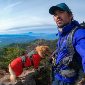 Imagem de um guia de trilhas e montanhismo vestindo jaqueta azul, boné azul e mochila. Está acompanhado de seu cão, um Golden Retriever, usando roupa vermelha e equipamento de segurança. Eles estão no alto de uma montanha. sobre mim, Daniel Tamaki, guia de montanhismo, Escalada Trilha Backcountry no Monte Fuji e Japão